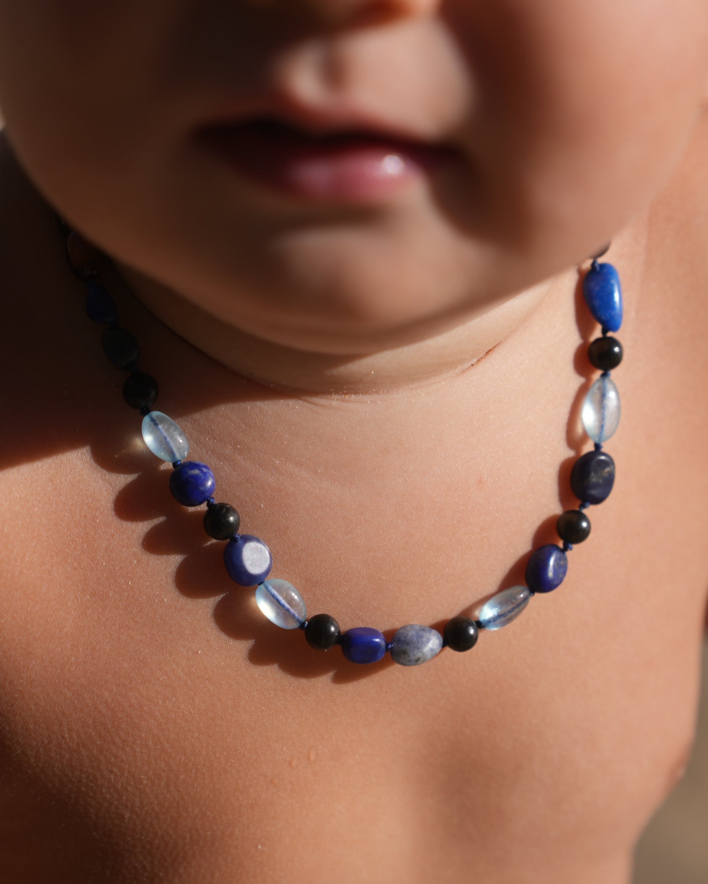 A close-up of a baby's chest and mouth area wearing a beaded shungite necklace with blue, black, and clear beads,that are Aquamarine, Lapis Lazuli, Sodaliteagainst a warm beige background.