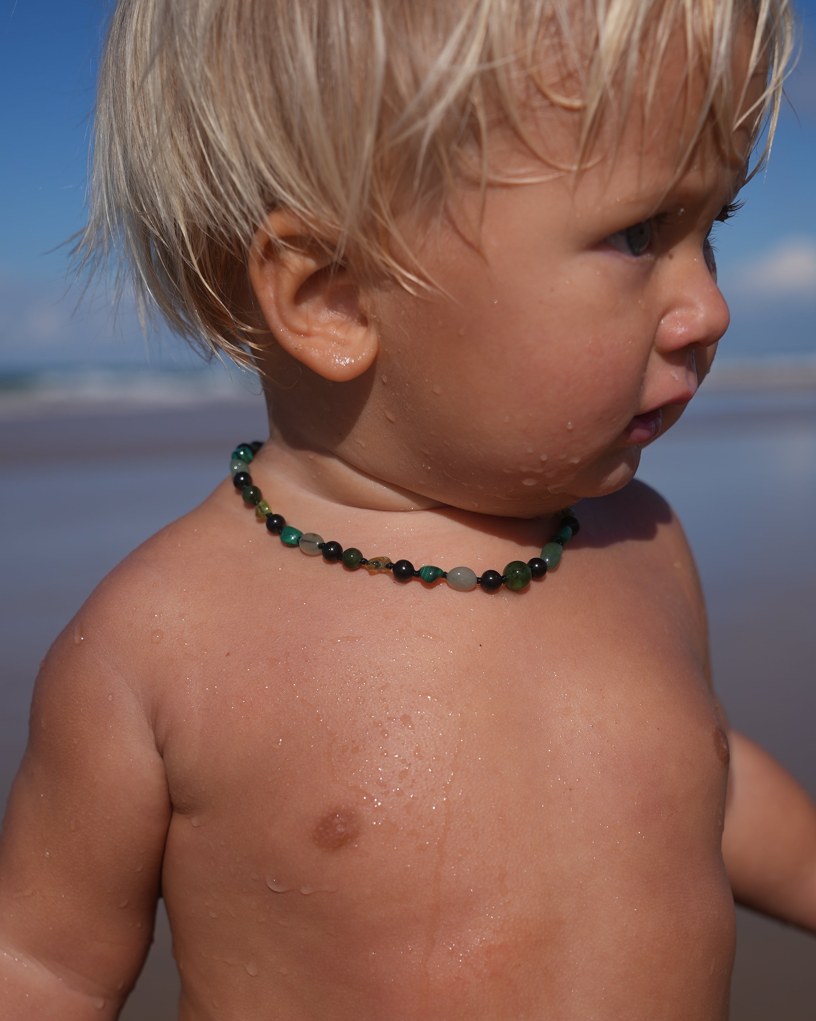 A close-up side profile of a young toddler with blonde hair standing at the beach, wearing a dark green beaded necklace.