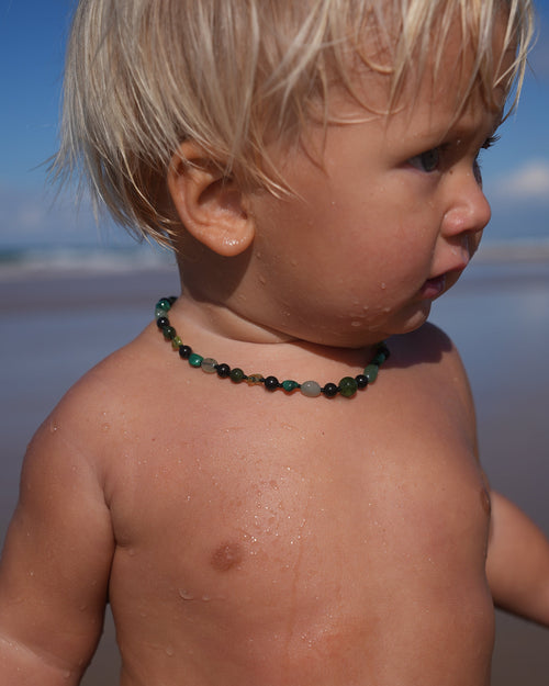 A close-up side profile of a young toddler with blonde hair standing at the beach, wearing a dark green beaded necklace.