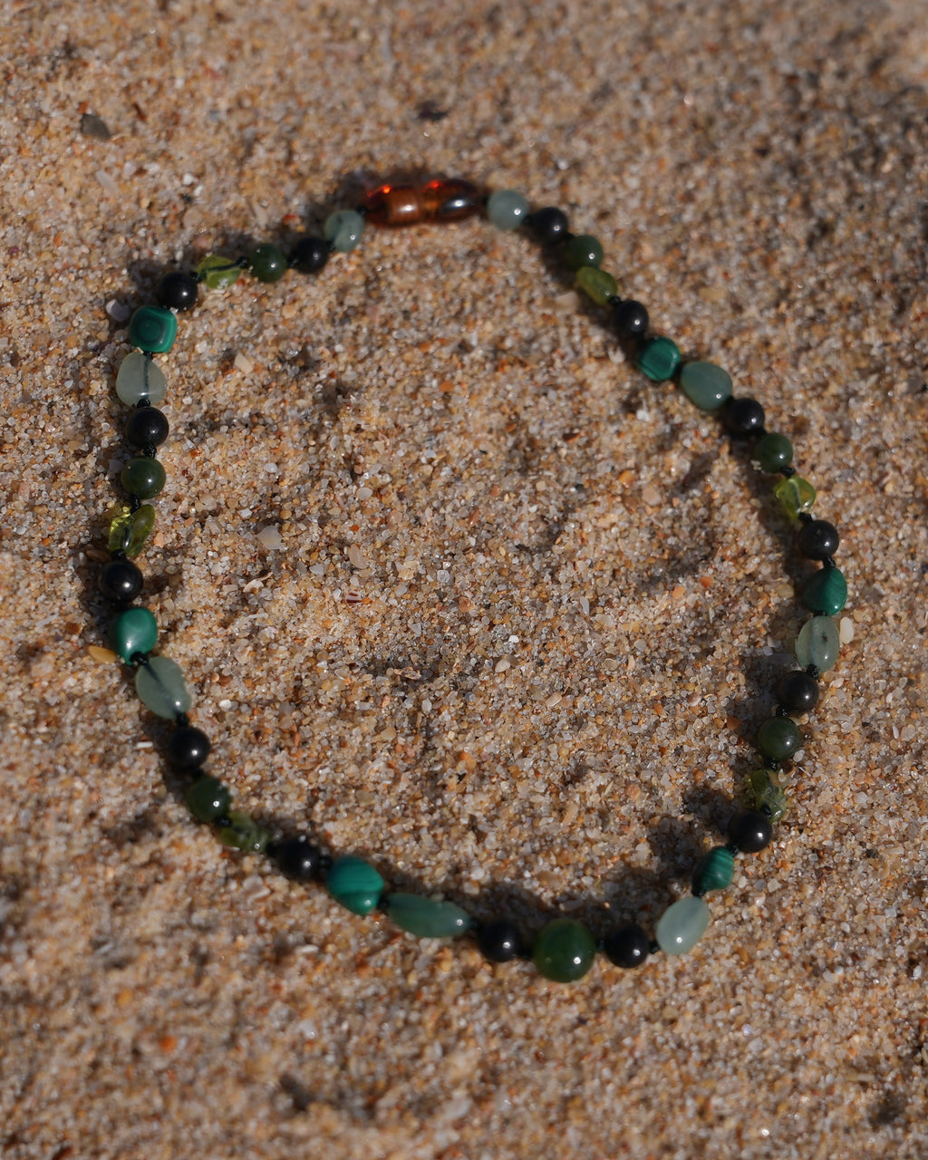 A beaded bracelet made of dark green, teal, and black beads with a small brown accent bead, arranged in a circle on a sandy surface.