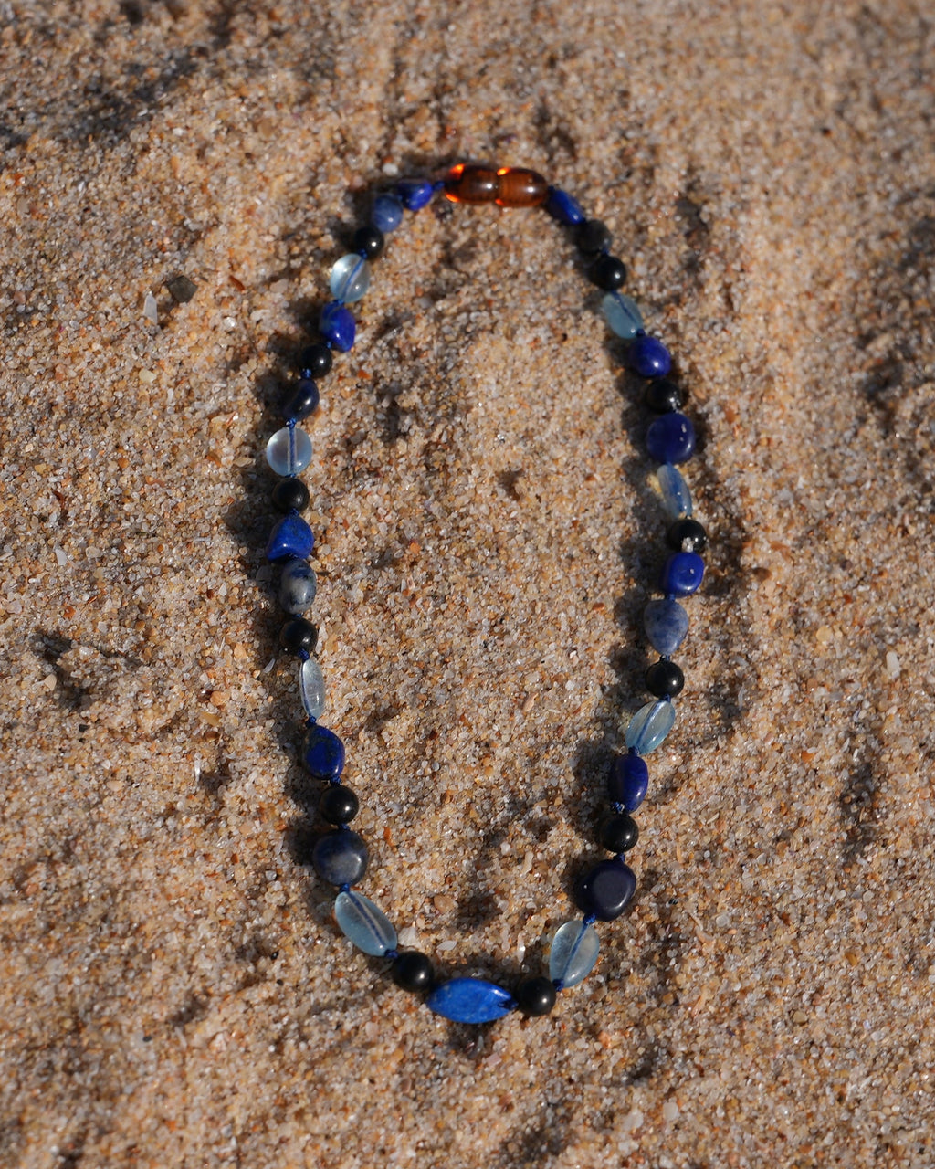 A beaded shungite necklace made of blue, navy, black, and light blue beads, which are Aquamarine, Lapis Lazuli, Sodalite and shungite arranged in an oval shape on a sandy beach.