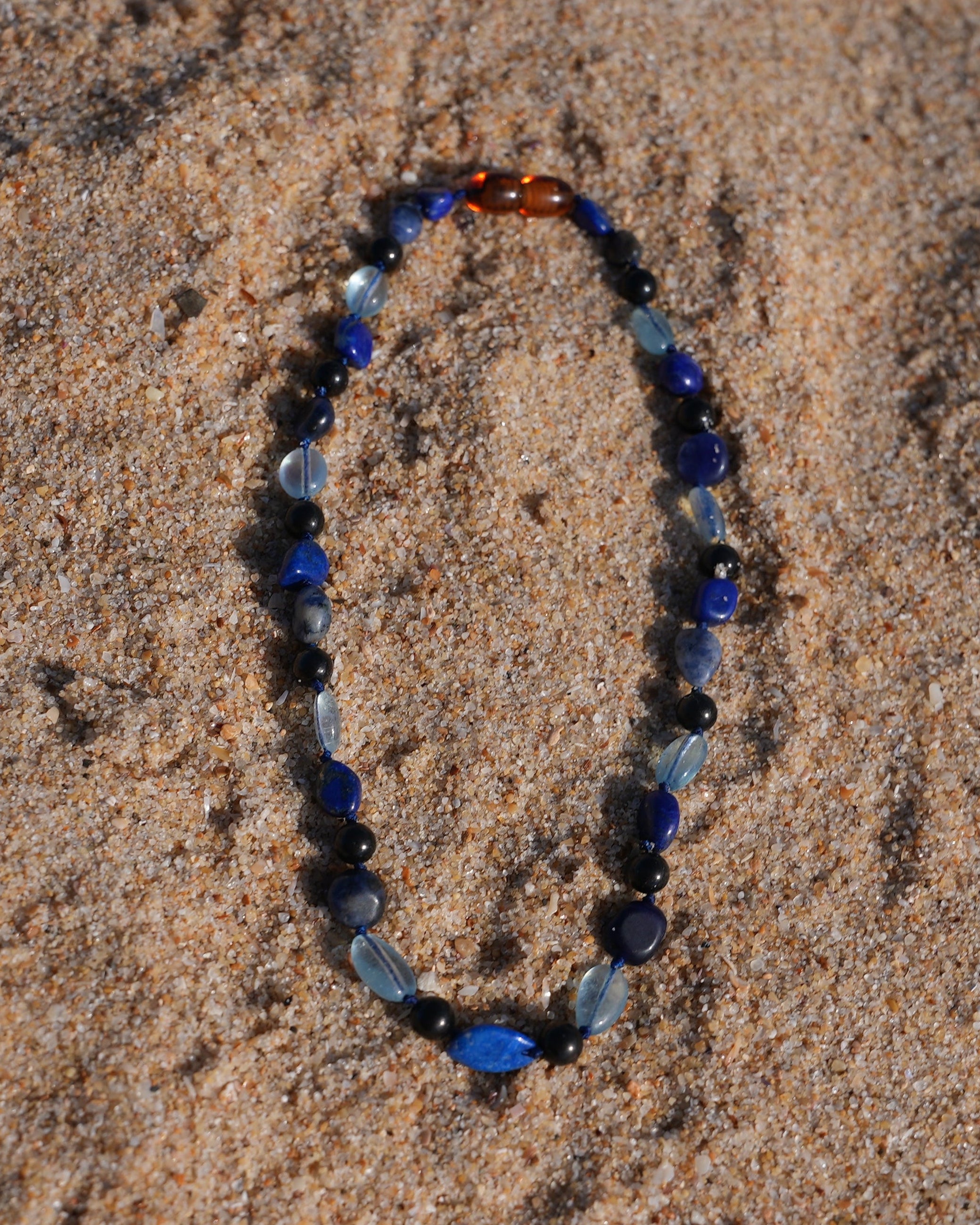 A beaded shungite necklace made of blue, navy, black, and light blue beads, which are Aquamarine, Lapis Lazuli, Sodalite and shungite arranged in an oval shape on a sandy beach.