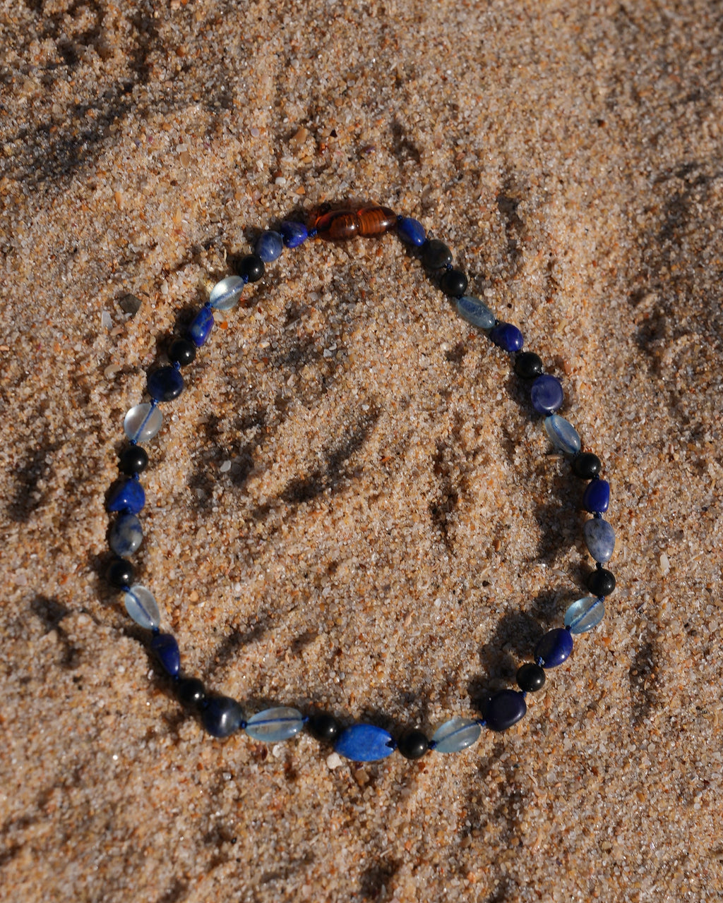 A beaded shungite necklace made of blue, navy, black, and light blue beads, which are Aquamarine, Lapis Lazuli, Sodalite and shungite arranged in an circle shape on a sandy beach.