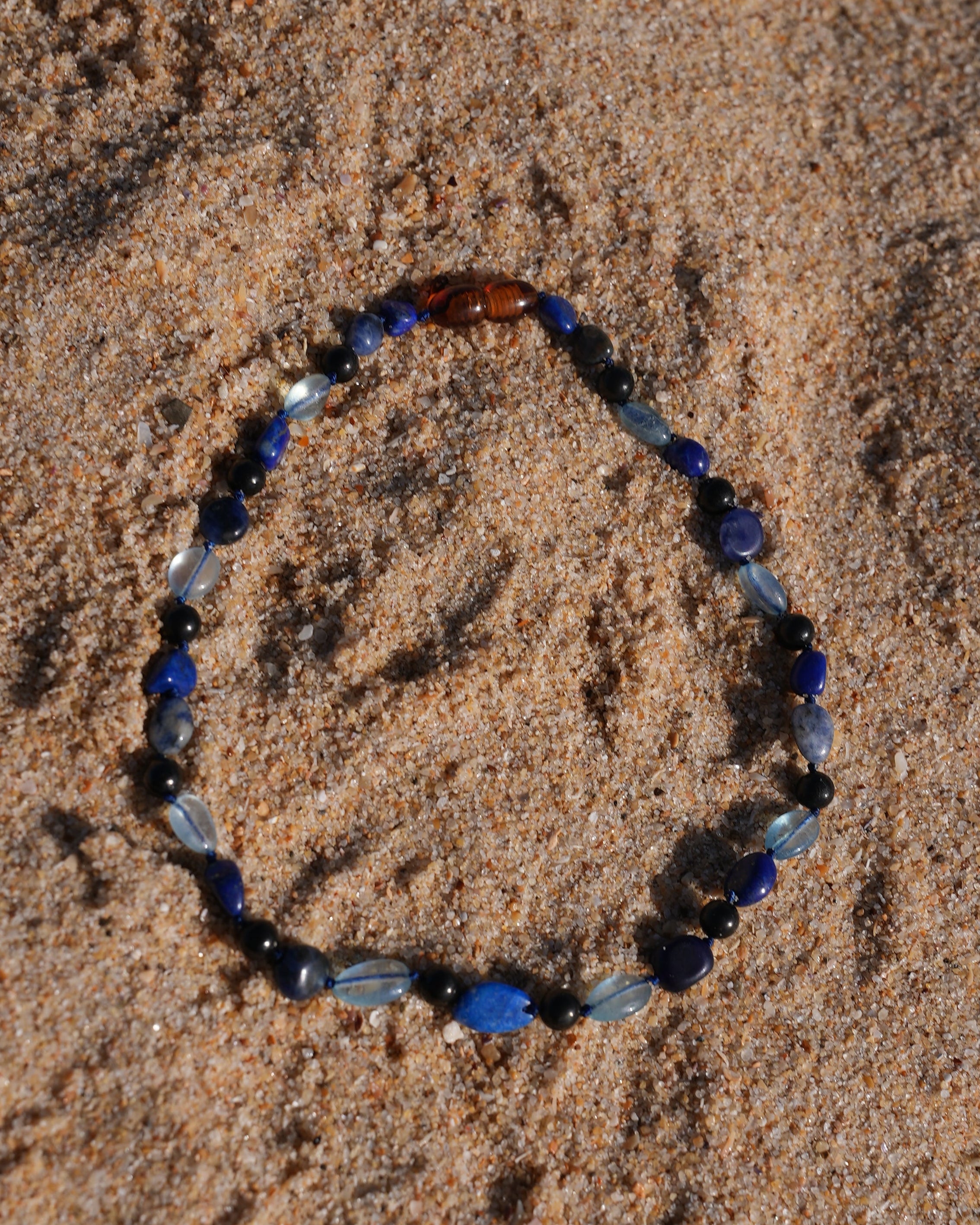 A beaded shungite necklace made of blue, navy, black, and light blue beads, which are Aquamarine, Lapis Lazuli, Sodalite and shungite arranged in an circle shape on a sandy beach.