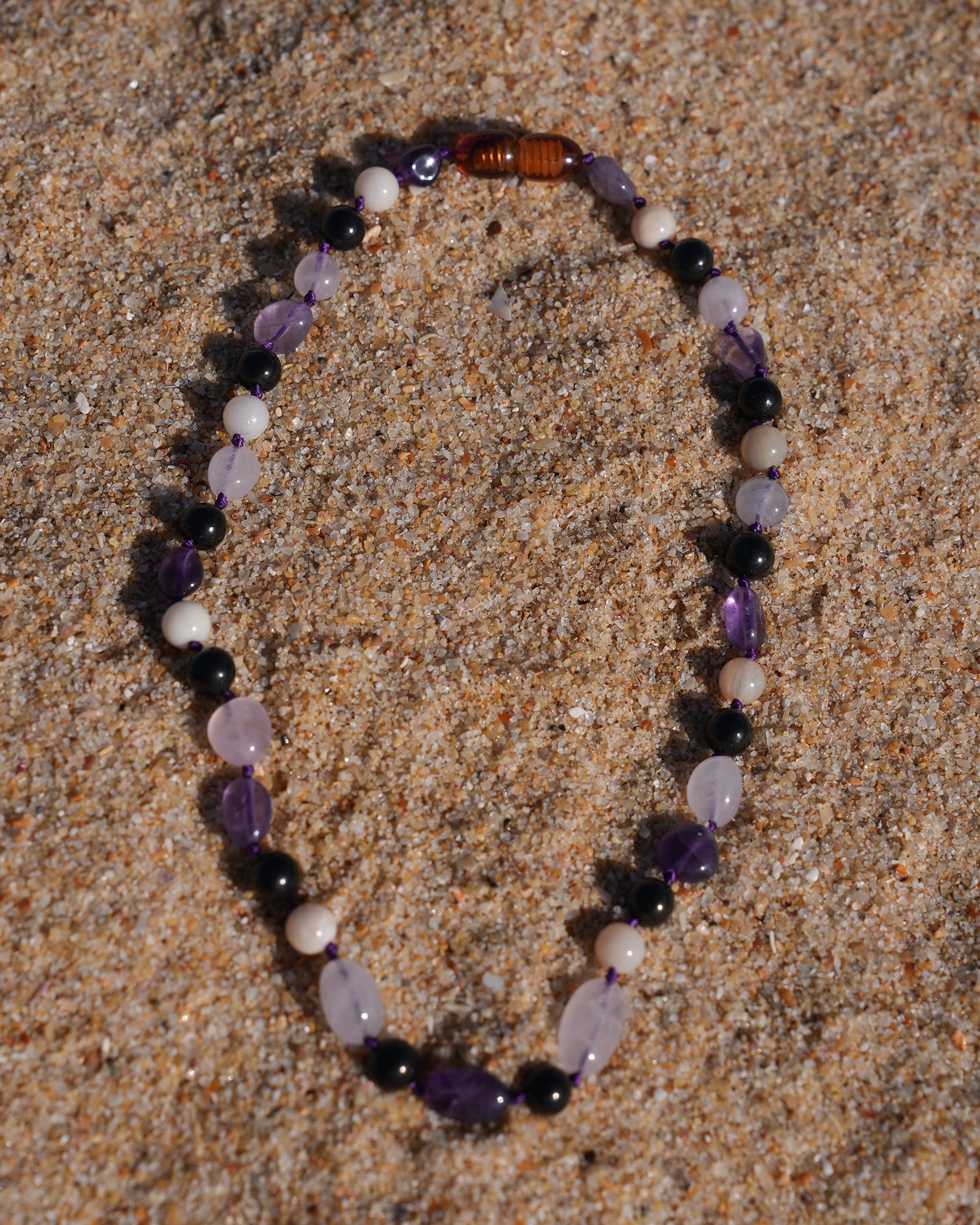 A beaded bracelet made of small polished stones in purple, black, white, and amber-brown, arranged in an oval on a sandy surface.