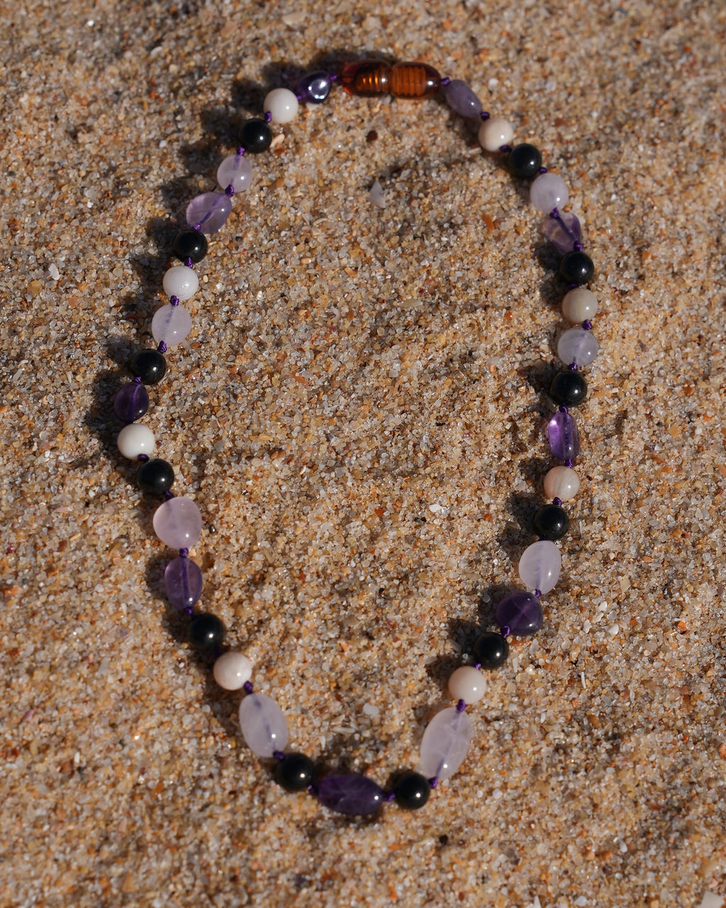A beaded necklace made of purple amethyst, white pearl-like beads, and a brown tiger's eye bead, arranged in an oval on a sandy surface.