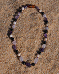 A beaded necklace made of purple amethyst, white pearl-like beads, and a brown tiger's eye bead, arranged in an oval on a sandy surface.