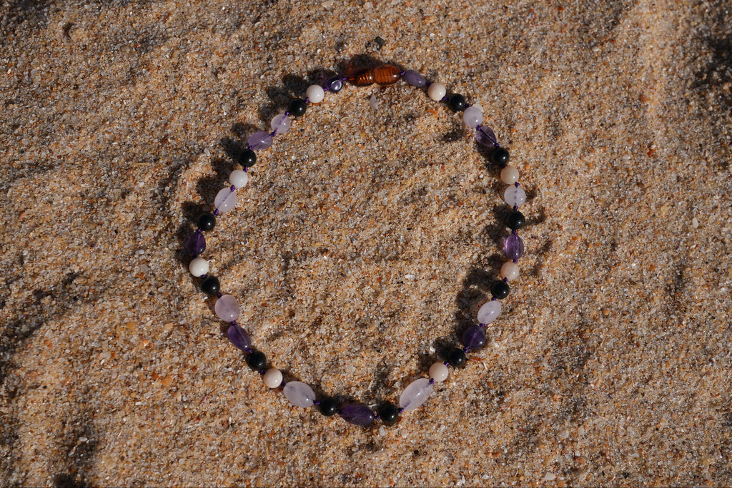 A beaded baby shungite necklace made of natural beach stones in purple, white, black, and amber, arranged in a circle on sand.