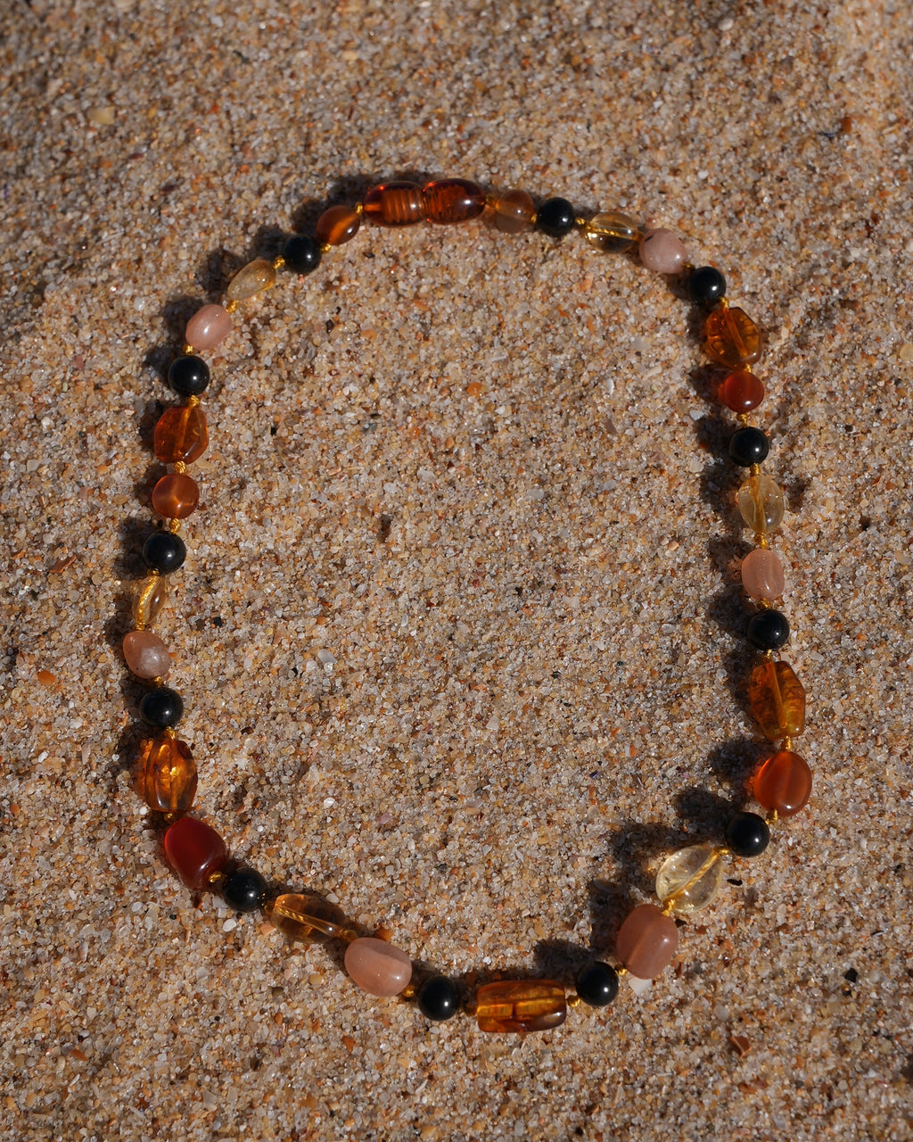 A beaded stretch bracelet made of mixed natural stones in amber, brown, pink, and black, arranged in a circle on a sandy beach.