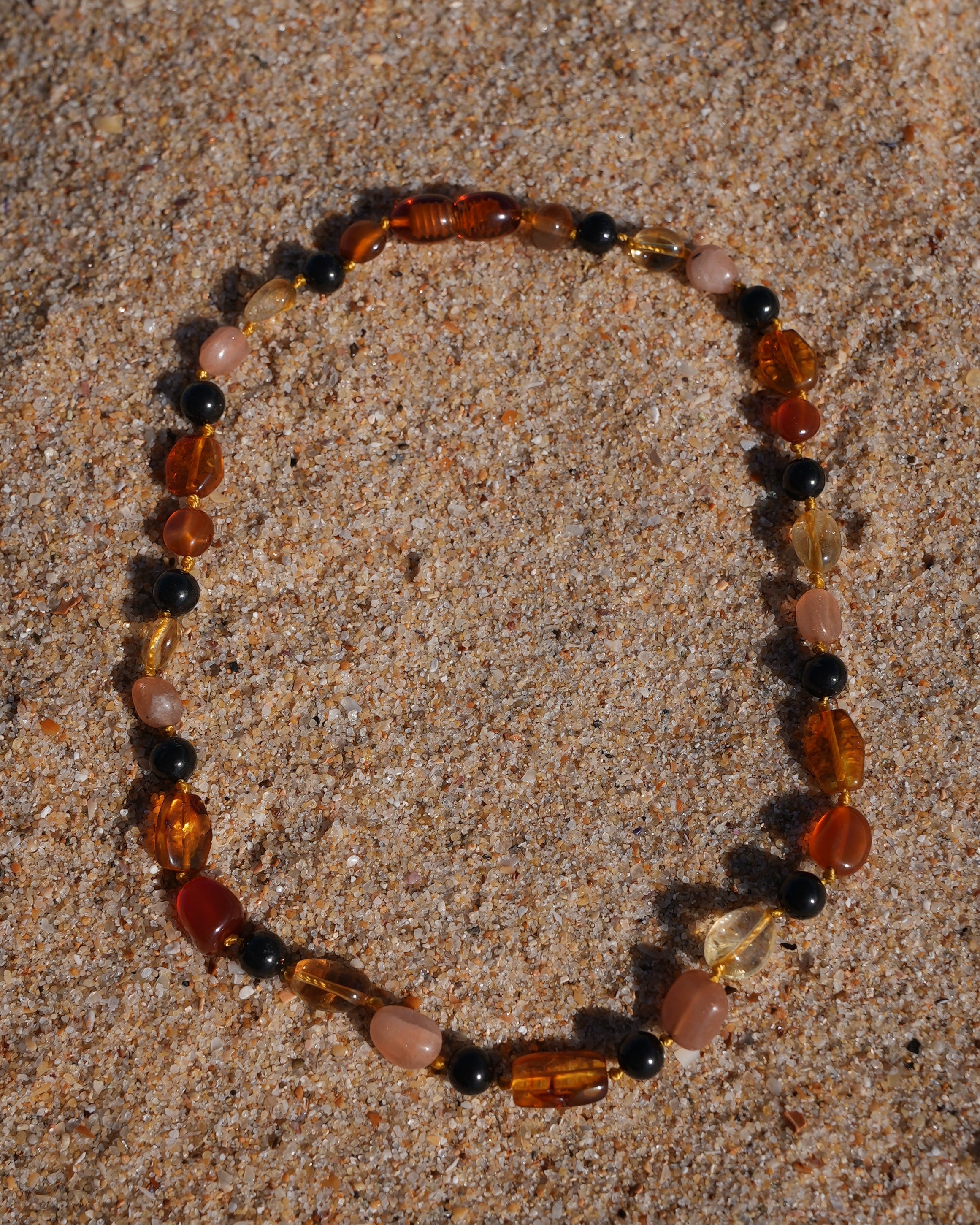 A beaded stretch bracelet made of mixed natural stones in amber, brown, pink, and black, arranged in a circle on a sandy beach.