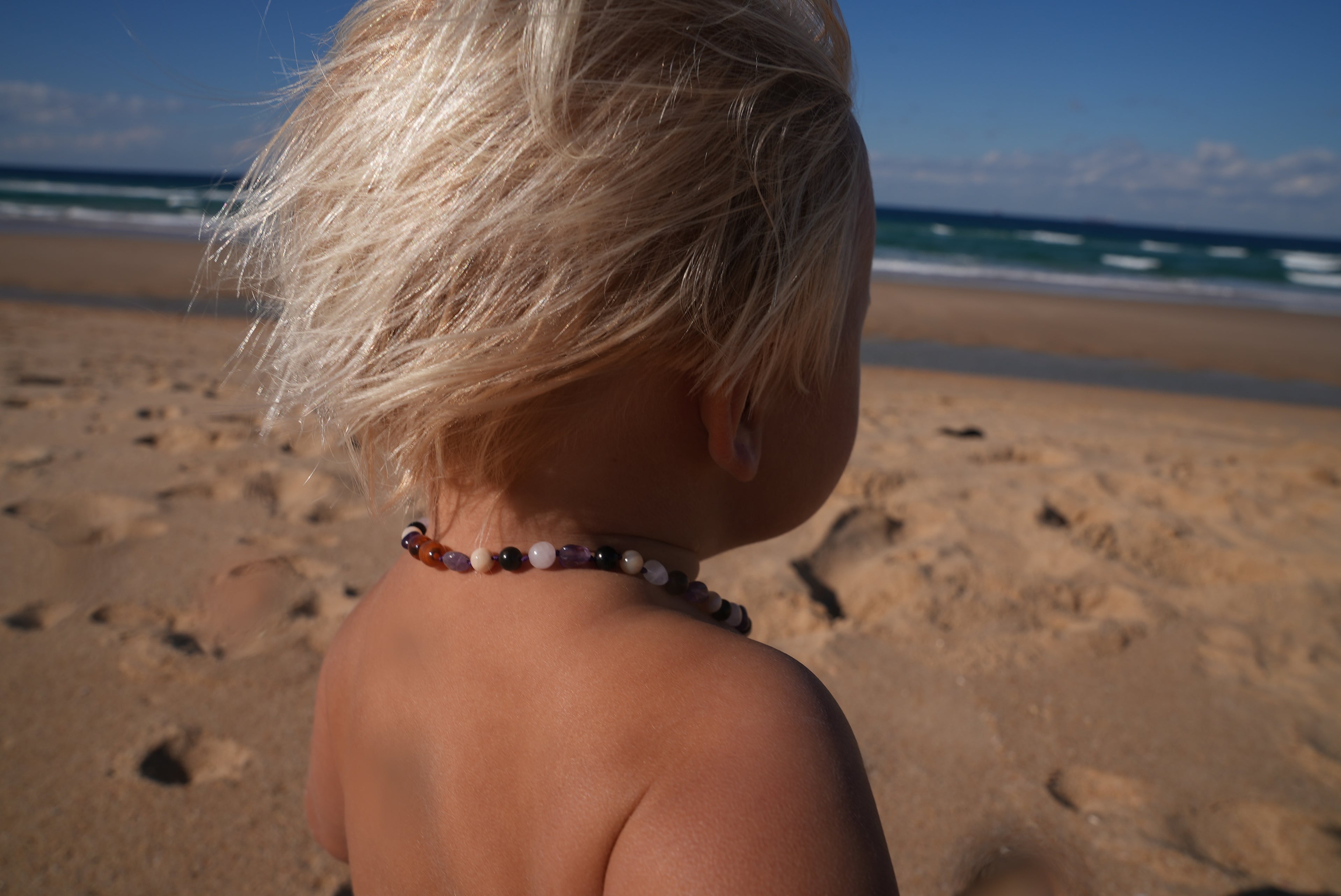 A close-up back view of a child with blonde hair on a sandy beach, wearing a beaded necklace with brown, white, and black beads.