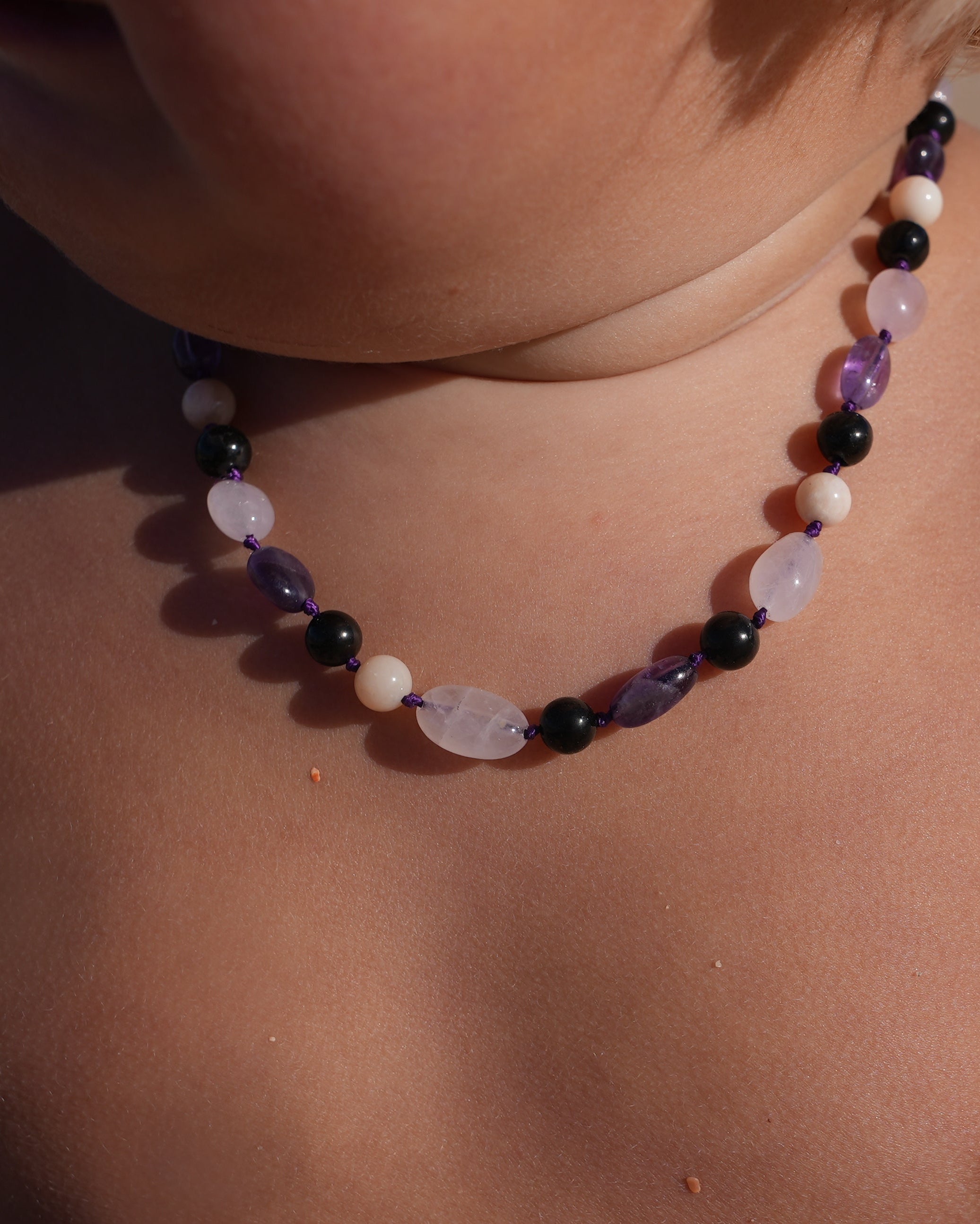 A close-up of a child's bare chest and neck at a beach, wearing a beaded necklace with purple, black, and white stones.