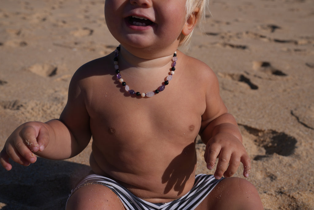 A close-up of a baby sitting on a sandy beach, wearing a colorful beaded necklace and black-and-white striped swim trunks.