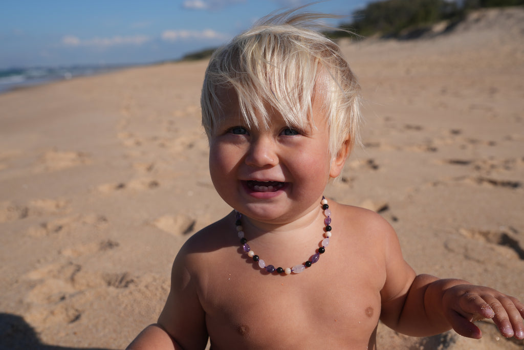A smiling toddler standing on a sandy beach wearing a colorful beaded necklace.