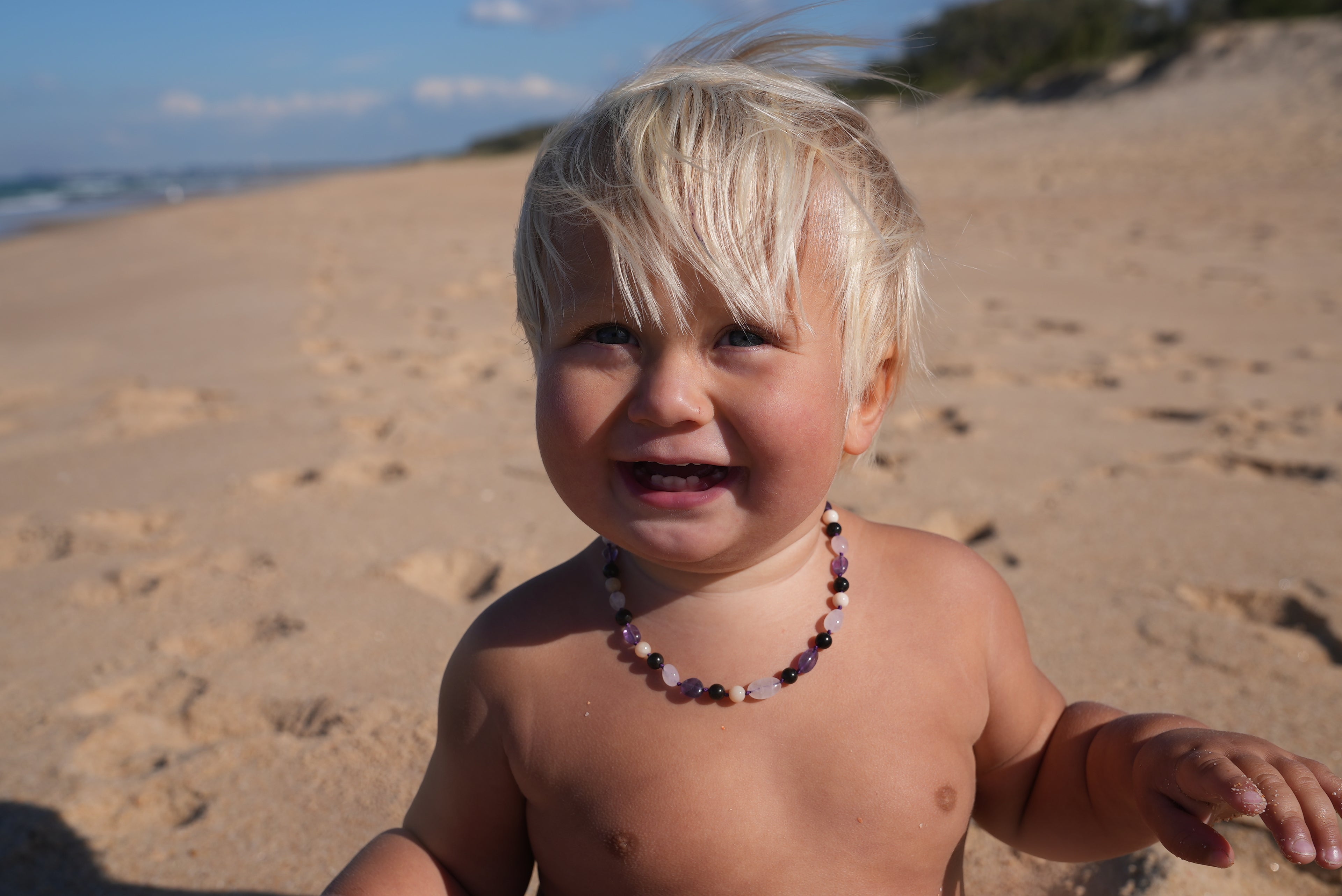 A smiling toddler standing on a sandy beach wearing a colorful beaded necklace.
