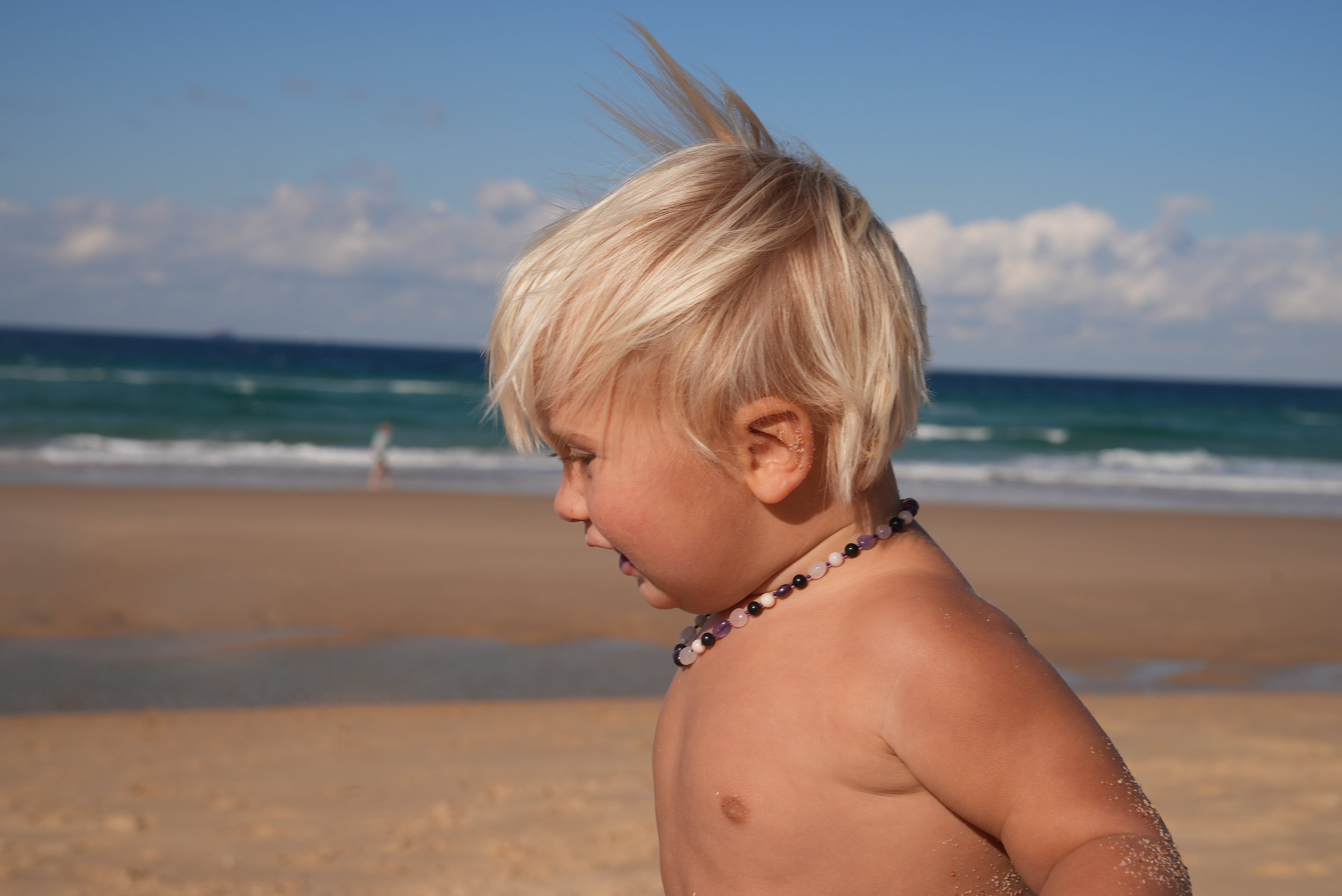 A young blonde child standing on a sandy beach with the ocean and blue sky in the background, wearing a beaded necklace.