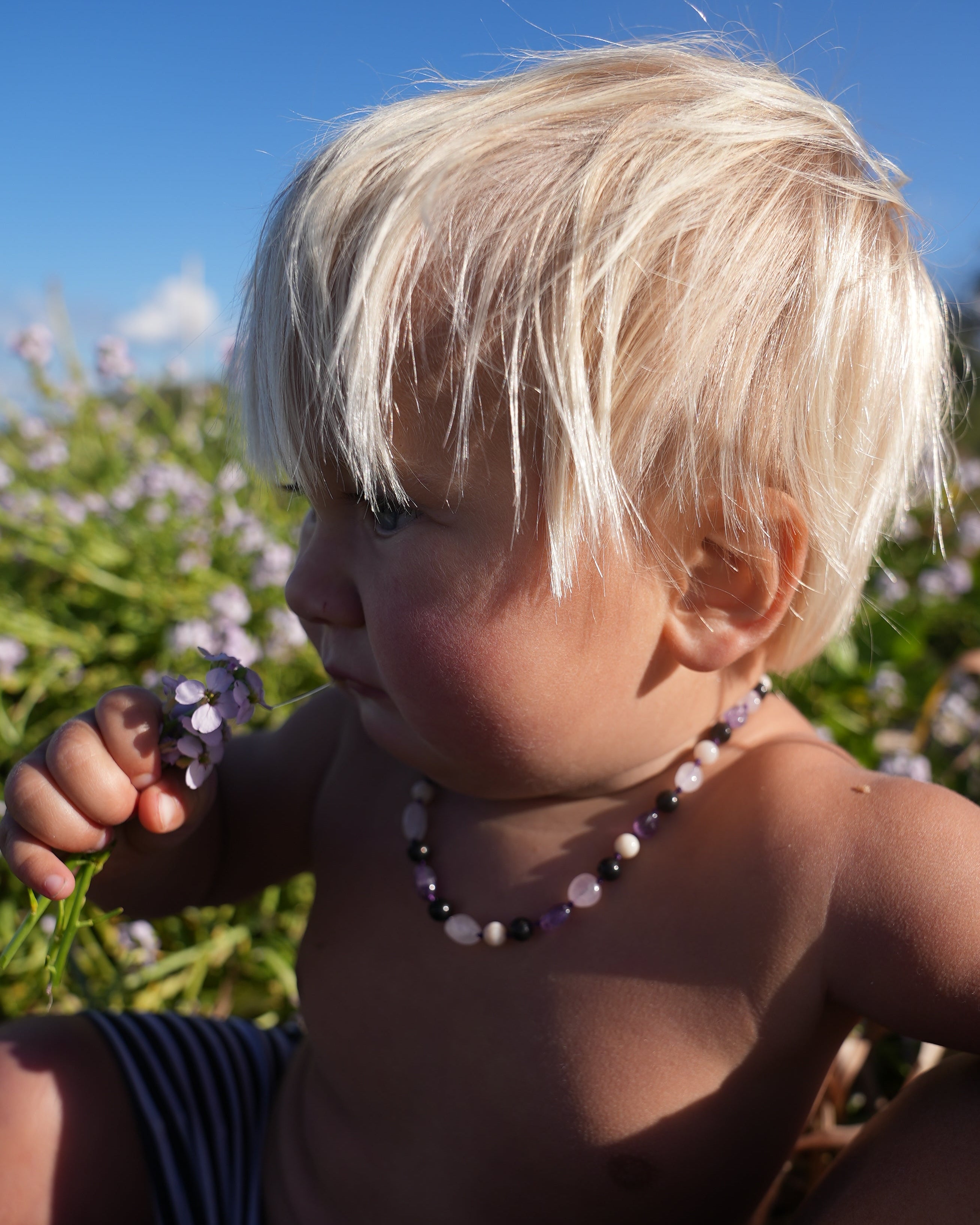 A close-up outdoor shot of a baby wearing a beaded necklace with purple and white beads, lying on grass with blue sky in the background.