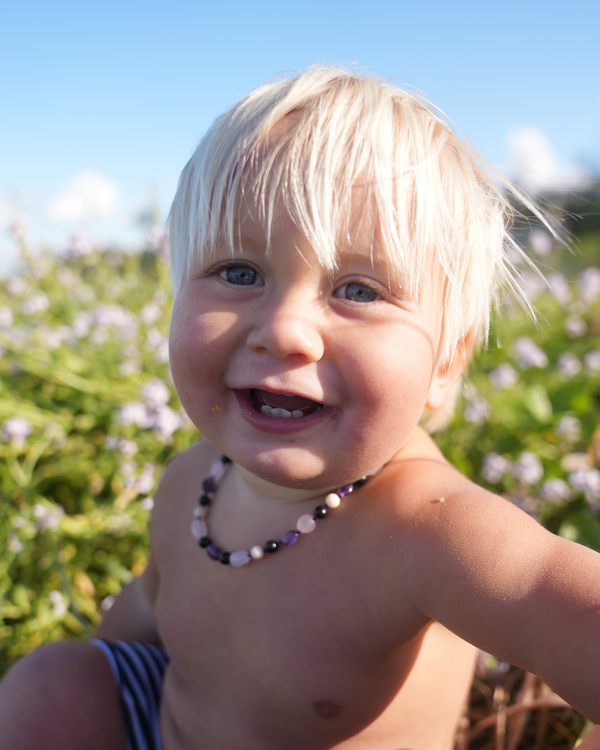 A close-up outdoor photo of a smiling young child lying on grass, wearing a beaded necklace and blue-and-white striped bottoms. The background shows green foliage and blue sky.