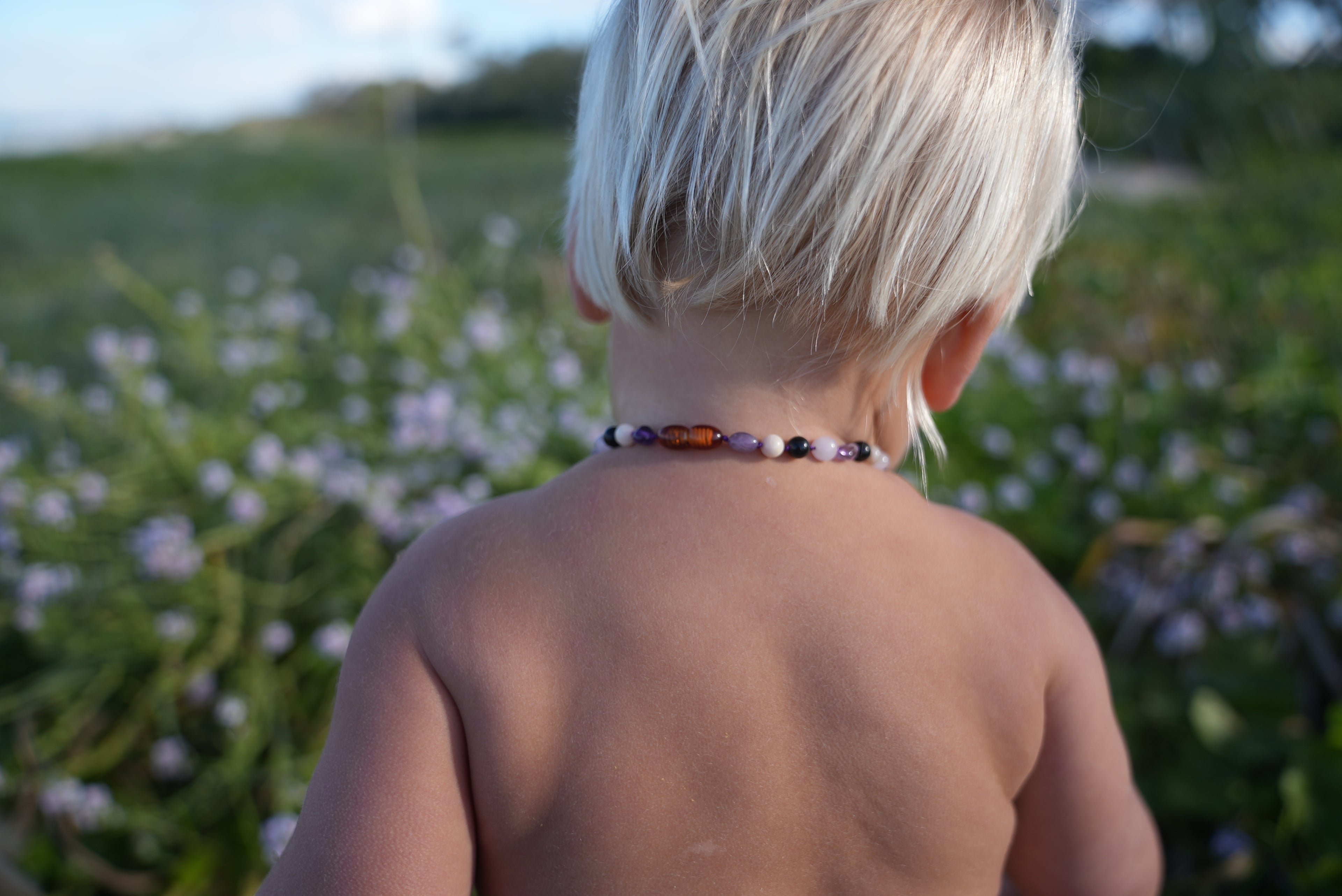 A close-up back view of a young child outdoors wearing a beaded necklace with purple, orange, and black beads, set against a soft-focus background of green foliage and small purple flowers.