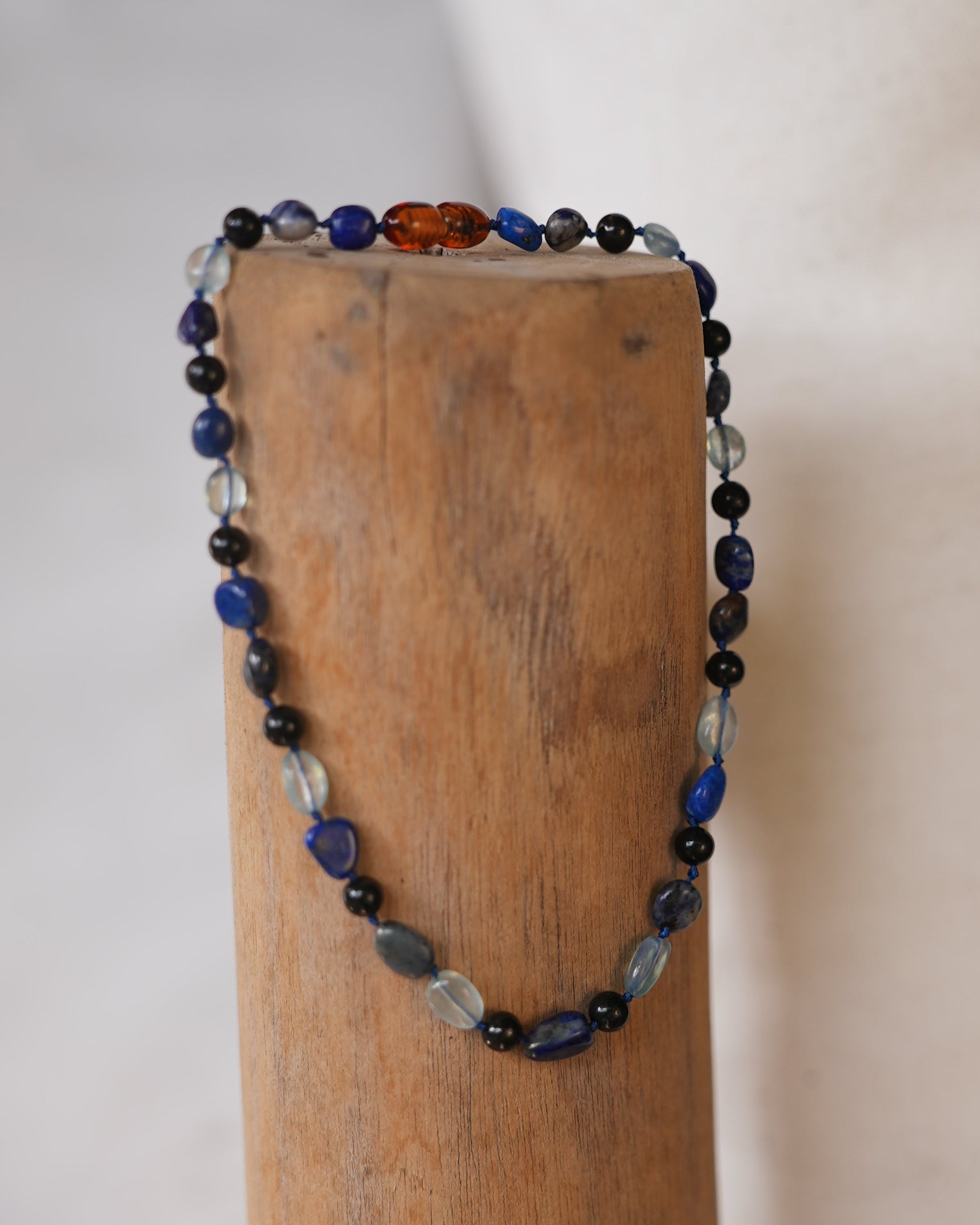 A beaded necklace with blue, black, and clear faceted beads draped over a wooden block against a light beige background.