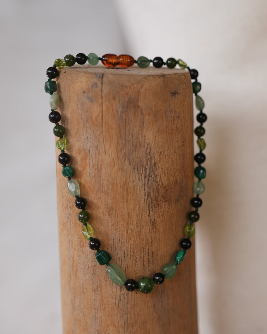 A beaded necklace with green, black, and amber-orange beads draped over a wooden block against a light neutral background.