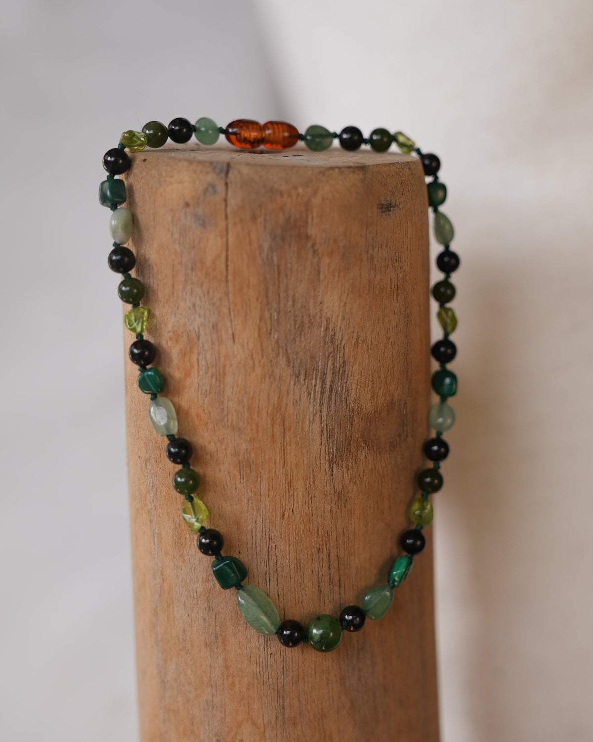 A beaded necklace with green, black, and amber-orange beads draped over a wooden block against a light neutral background.