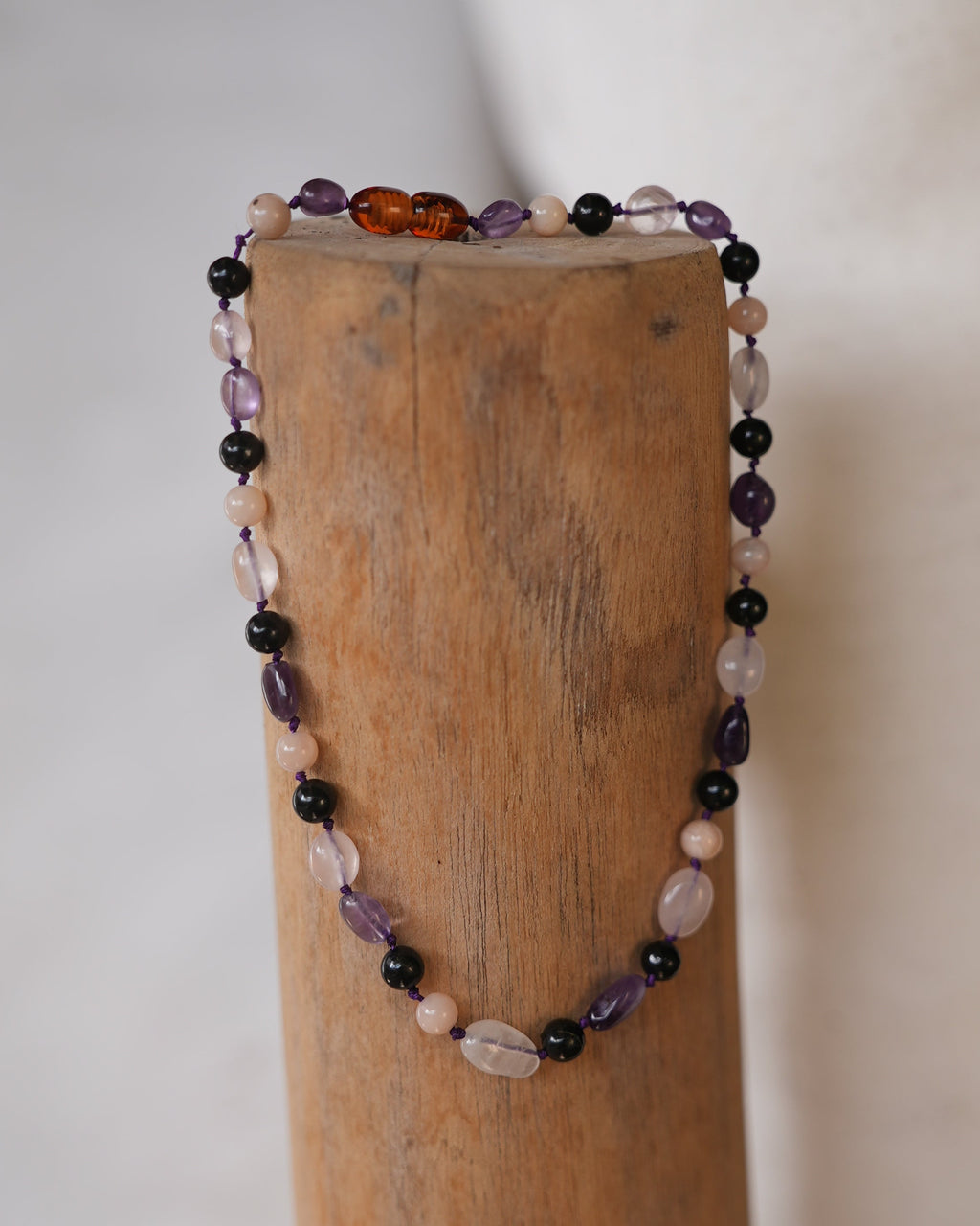 A beaded bracelet with mixed purple, black, pink, and amber beads draped over a wooden dowel against a light fabric background.