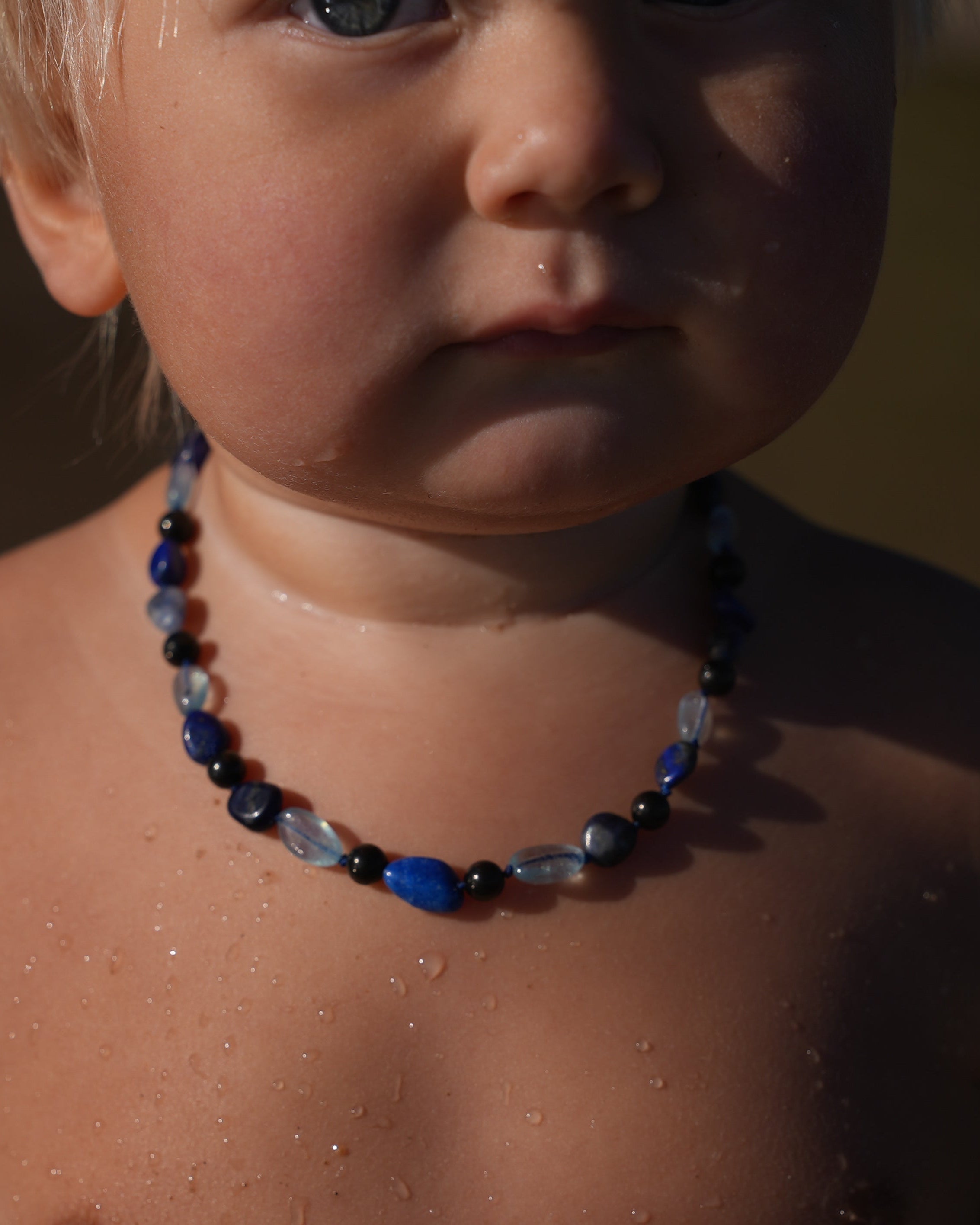 A close-up shot of a baby wearing a beaded shungite necklace with blue, black, and clear beads,that are Aquamarine, Lapis Lazuli, Sodalite set against a warm, blurred background.