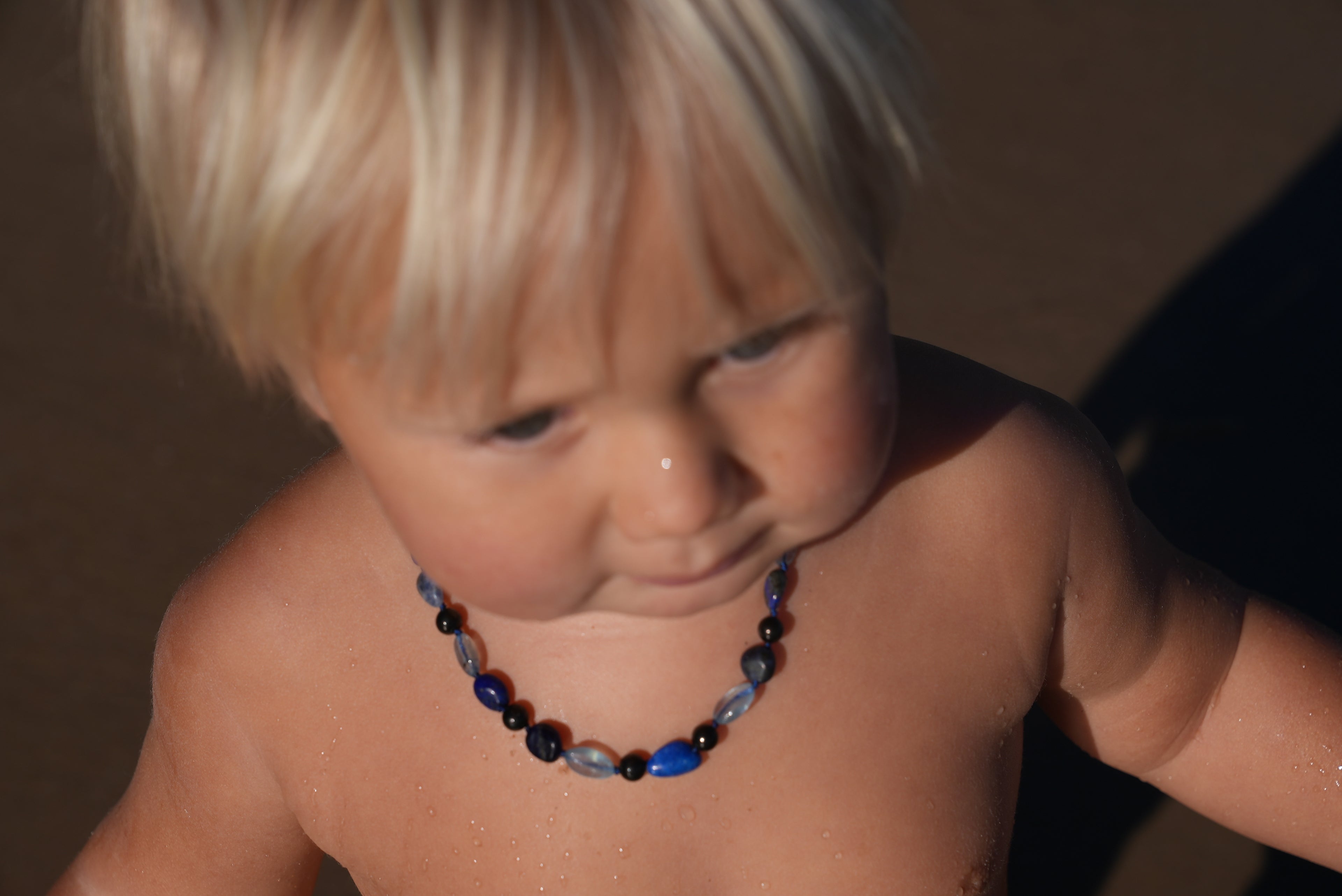 A close-up of a baby wearing a colorful beaded shungite necklace with blue, black, and clear beads,that are Aquamarine, Lapis Lazuli, Sodalite photographed in warm sunlight.