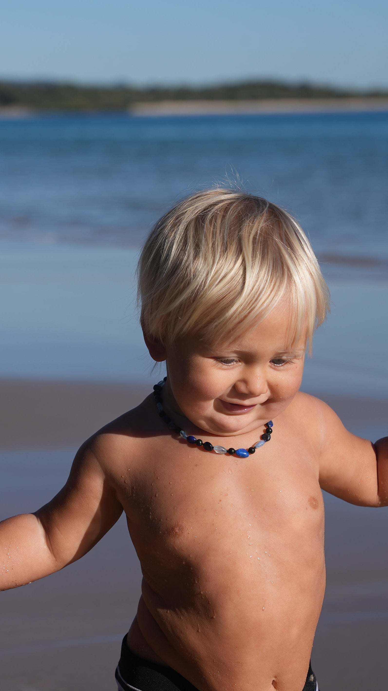 A young blonde toddler standing on a sandy beach with the ocean and distant shoreline in the background, smiling and looking to the side wearing a shungite necklace with blue, black, and clear beads,that are Aquamarine, Lapis Lazuli, Sodalite